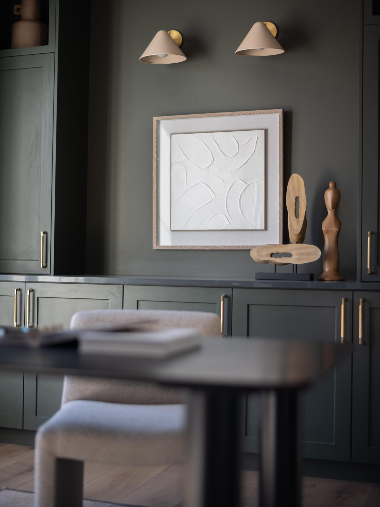 Sage green built-ins with brass hardware, a textured white canvas and carved wood sculptures under cone sconces
